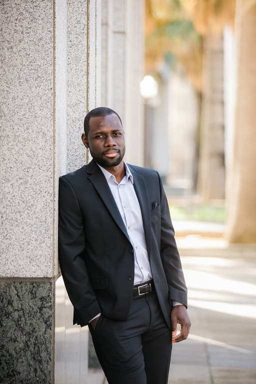 Stock photo of Fortune Vieyra in a black suit leaning against a stone wall with one hand in his pocket, used as the reference image