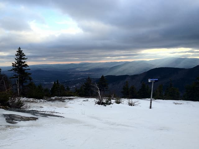 Test image: a snowy mountain summit with a trail sign and evergreens, overlooking distant ridges under heavy clouds
